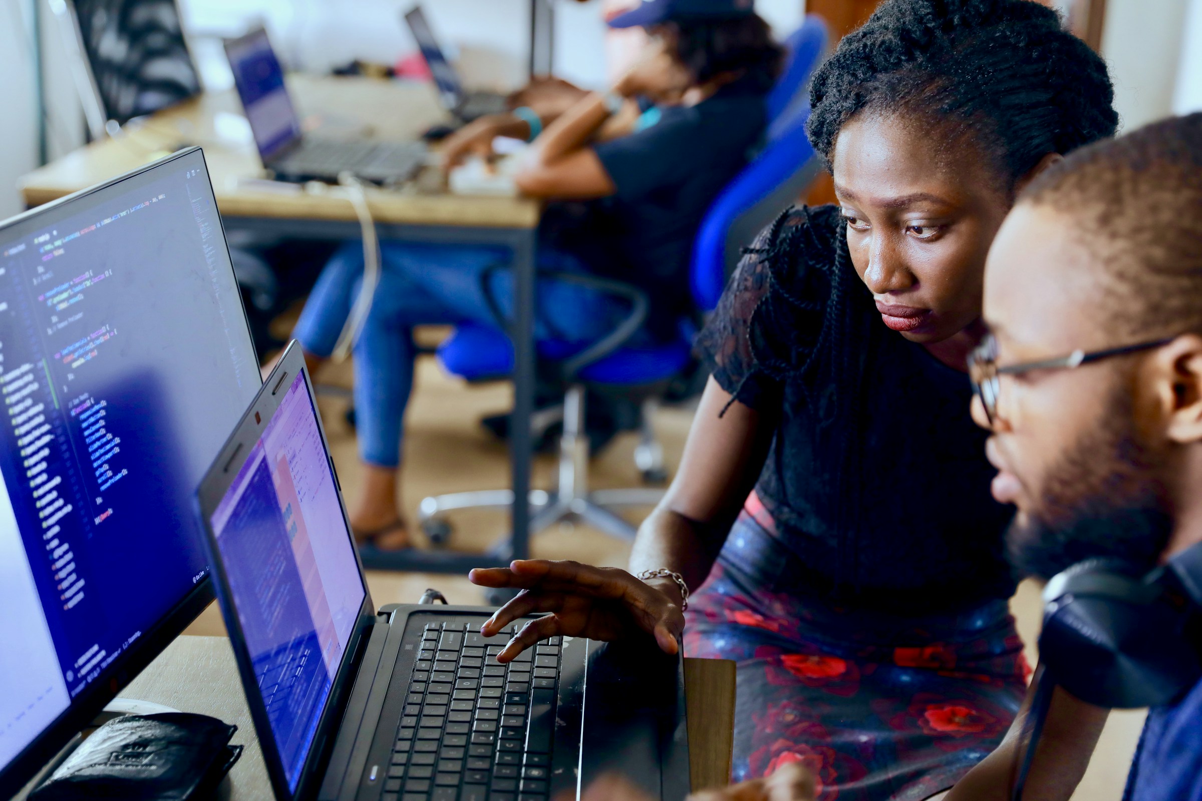 Two developers reviewing code together on a laptop in a busy workspace